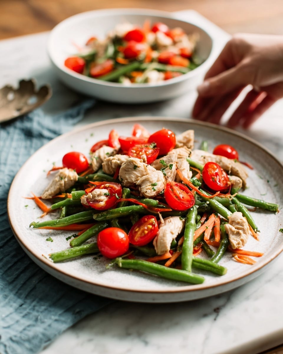 The image shows a white plate on a white marbled surface with a colorful dish. The dish has three layers: the bottom layer is a mix of cooked green beans and thin carrot strips, the middle layer consists of small cooked chicken pieces that are light brown with a slightly crispy texture, and the top layer is made of bright red cherry tomato halves scattered evenly over the chicken. A white bowl in the background holds the same dish, blurred slightly. The lighting is soft and natural, enhancing the fresh colors. A woman's hand is reaching toward the plate from the right side. Photo taken with an iphone --ar 4:5 --v 7