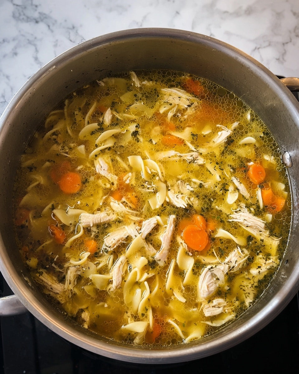 This image shows a large silver pot filled with chicken noodle soup. The soup has many layers visible: shredded light beige chicken pieces, orange carrot chunks, flat whitish noodles, and some green herbs all mixed in a golden yellow broth. The pot rests on a stove with a dark surface edge and the background features a white marbled texture. The soup looks hot and ready to serve, with small oil bubbles floating on the surface. Photo taken with an iphone --ar 4:5 --v 7