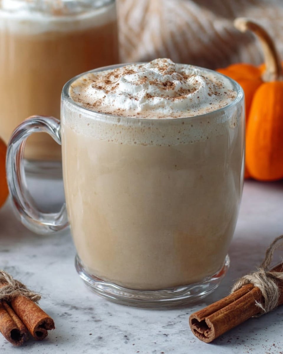 The image shows a clear glass mug filled with a creamy light beige drink. The surface of the drink is topped with a layer of white whipped cream sprinkled with fine brown powder, likely cinnamon or nutmeg. The mug has a thick handle on the left side and sits on a white marbled surface. In the background, a small orange pumpkin and another glass with a similar drink are softly visible. A bundle of cinnamon sticks tied with coarse string lies near the mug. Photo taken with an iphone --ar 4:5 --v 7