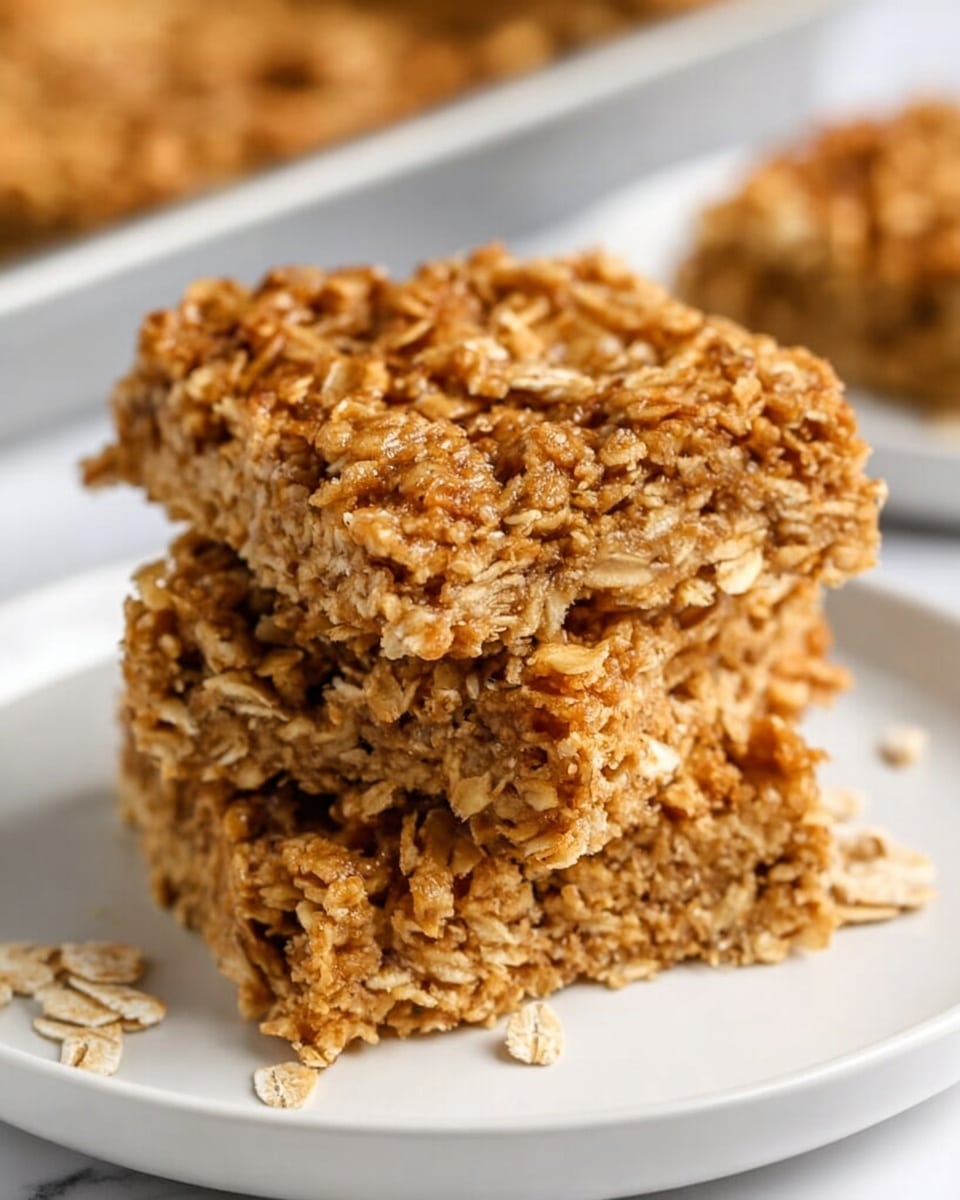 The image shows three square oat bars stacked on a white plate with a white marbled surface underneath. Each bar has a rough, crumbly texture with visible oat flakes coated in a golden brown mixture, giving them a slightly shiny appearance. Small clusters of oats stick out irregularly from the edges, and some loose oats lie scattered on the plate near the bars. The background is softly blurred, hinting at more oat bars on trays. photo taken with an iphone --ar 4:5 --v 7