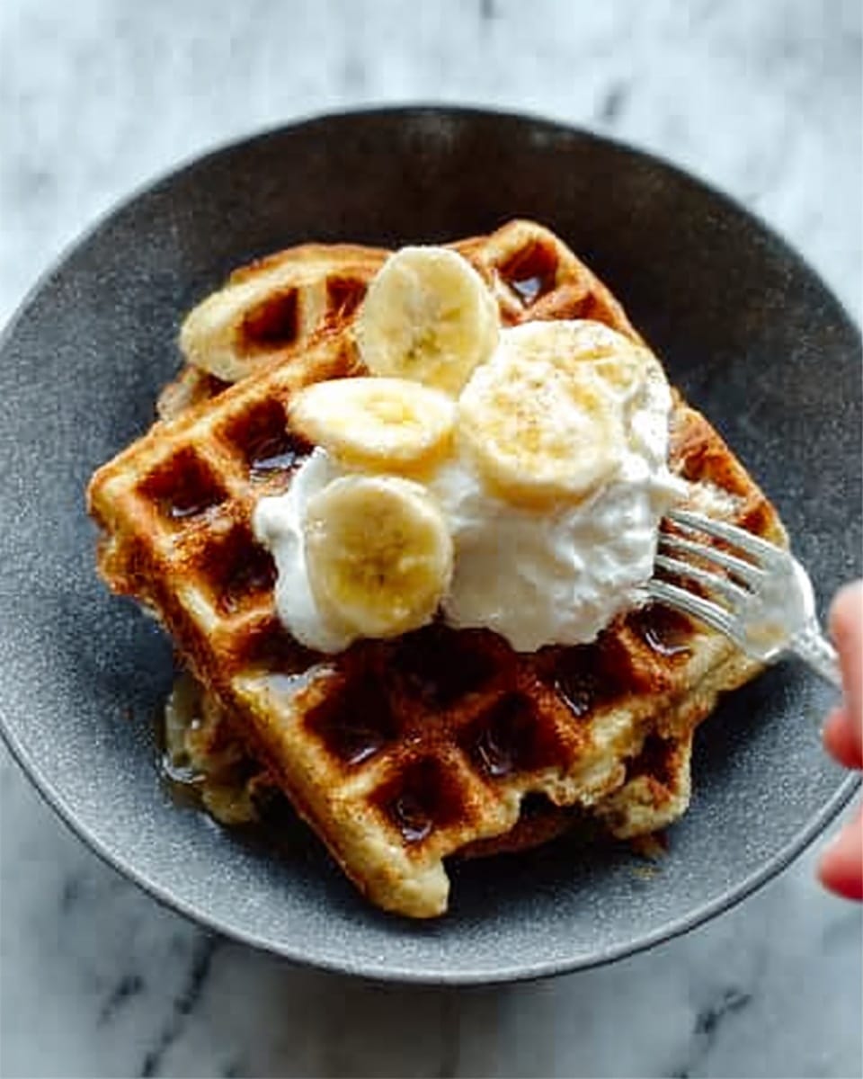 A close-up of a dark gray bowl with a stack of two waffles inside it; the waffles are golden brown with a crispy texture and a grid pattern. On top, there are three banana slices placed in a triangle shape along with two dollops of whipped cream that look soft and fluffy. A drizzle of syrup adds a shiny, sticky layer over the waffles, and a silver fork with a woman's hand holding it is touching one side of the waffle stack. The bowl is placed on a white marbled surface. photo taken with an iphone --ar 4:5 --v 7