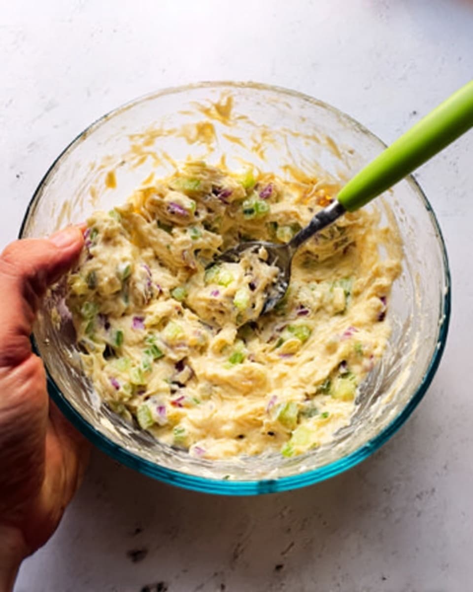 This image shows a clear glass bowl filled with a creamy mixture. The mixture has a pale yellow base with visible pieces of finely chopped green celery and small bits of purple onion spread throughout. The texture appears smooth with tiny chunks, and there is a metal spoon with a green handle resting inside the bowl. A woman's hand is holding the bowl from below. The background features a soft light source, and everything is seen on a white marbled surface. photo taken with an iphone --ar 4:5 --v 7