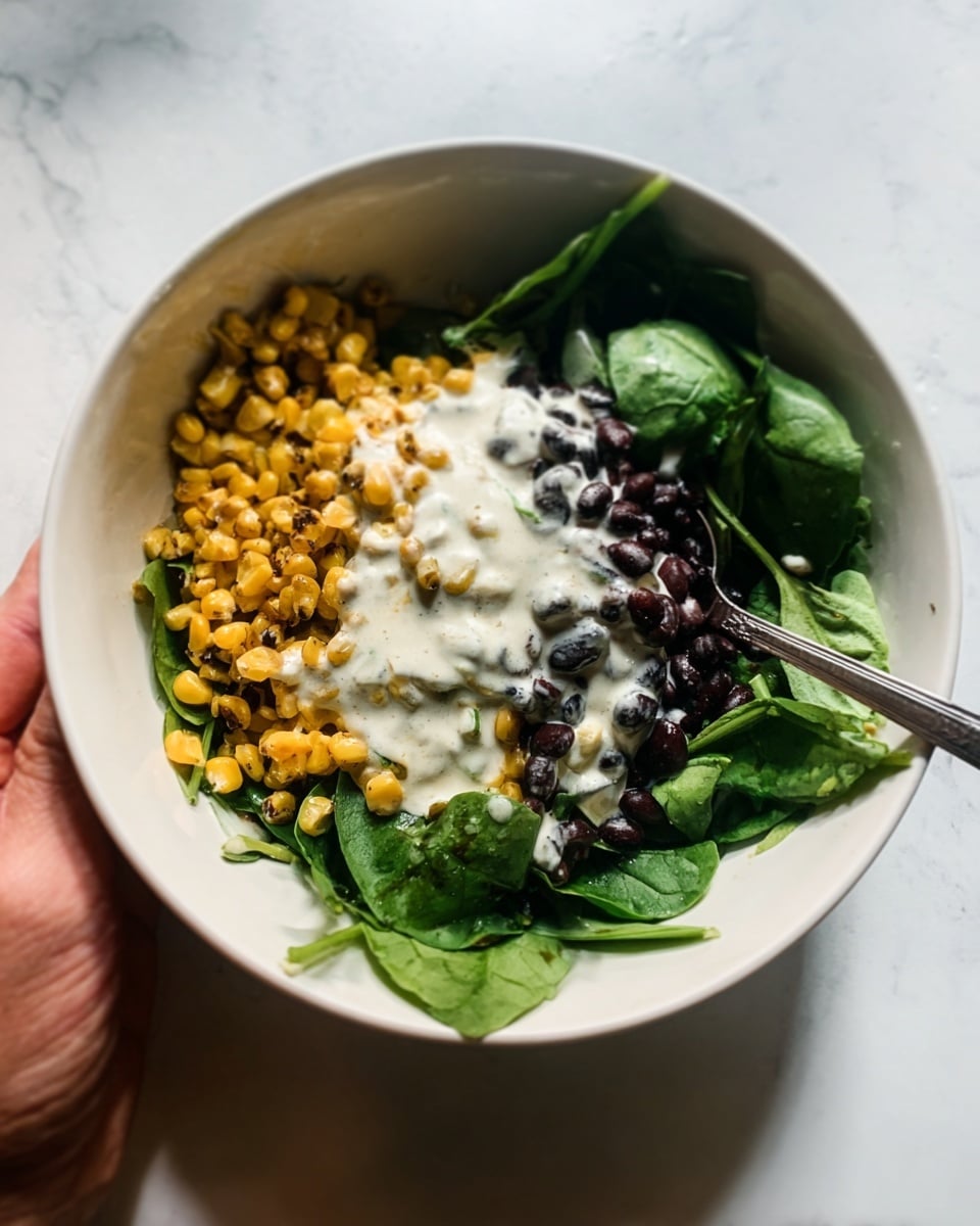A white bowl sits on a white marbled surface, filled with three main layers: at the bottom right, there are fresh dark green spinach leaves; to the left, a layer of golden-yellow roasted corn kernels; and at the top right, a pile of small black beans. On top of these layers, a creamy white sauce with a smooth texture is drizzled, slightly blending with the spinach and beans. A white spoon is placed inside the bowl, angled towards the viewer, and a woman's hand holding the bowl is partly visible from the left side. Photo taken with an iphone --ar 4:5 --v 7
