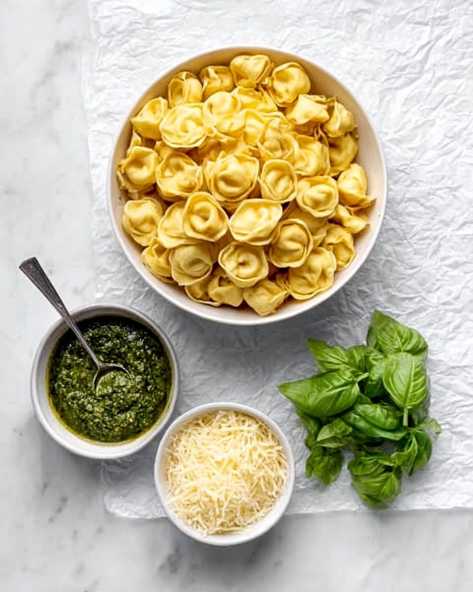 A round white bowl filled with a large amount of golden-yellow tortellini pasta sits at the top center on a white crumpled paper texture. Below it, slightly left, is a small white bowl with dark green pesto sauce and a small silver spoon inside. To the right of the pesto bowl, there is a small white bowl filled with finely shredded pale yellow cheese. Next to the cheese bowl is a small bunch of fresh, bright green basil leaves. The whole scene is set on a white marbled surface. photo taken with an iphone --ar 4:5 --v 7