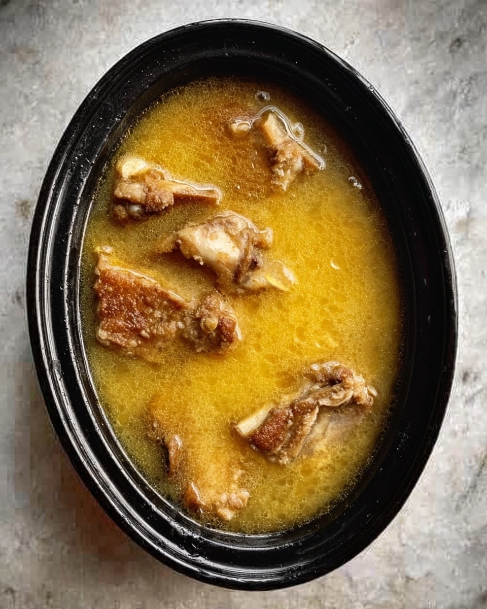 The image shows a black oval dish filled with a golden yellow broth, which has a slightly thick texture. Inside the broth, there are several pieces of cooked meat with visible bones, evenly spaced throughout the dish. The surface beneath the dish is a white marbled texture. The photo taken with an iphone --ar 4:5 --v 7