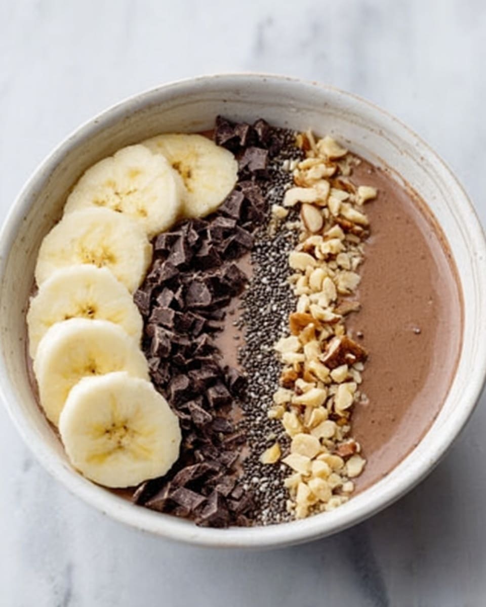 A white bowl filled with a smooth light brown chocolate smoothie base is decorated on top with three main sections: on the left side, broken dark chocolate sandwich cookies with white cream filling are placed in a rough pile; on the right side, two round chocolate candies with a nut center are placed side by side; on the top right, a row of thin light yellow banana slices is neatly arranged. Scattered on top are small dark chocolate chips and chunks of milk chocolate squares, all drizzled with a glossy dark chocolate sauce. The bowl is set on a white marbled surface with a gray and white striped cloth nearby alongside whole and broken cookies, chocolate chips, and a piece of milk chocolate. Photo taken with an iphone --ar 4:5 --v 7