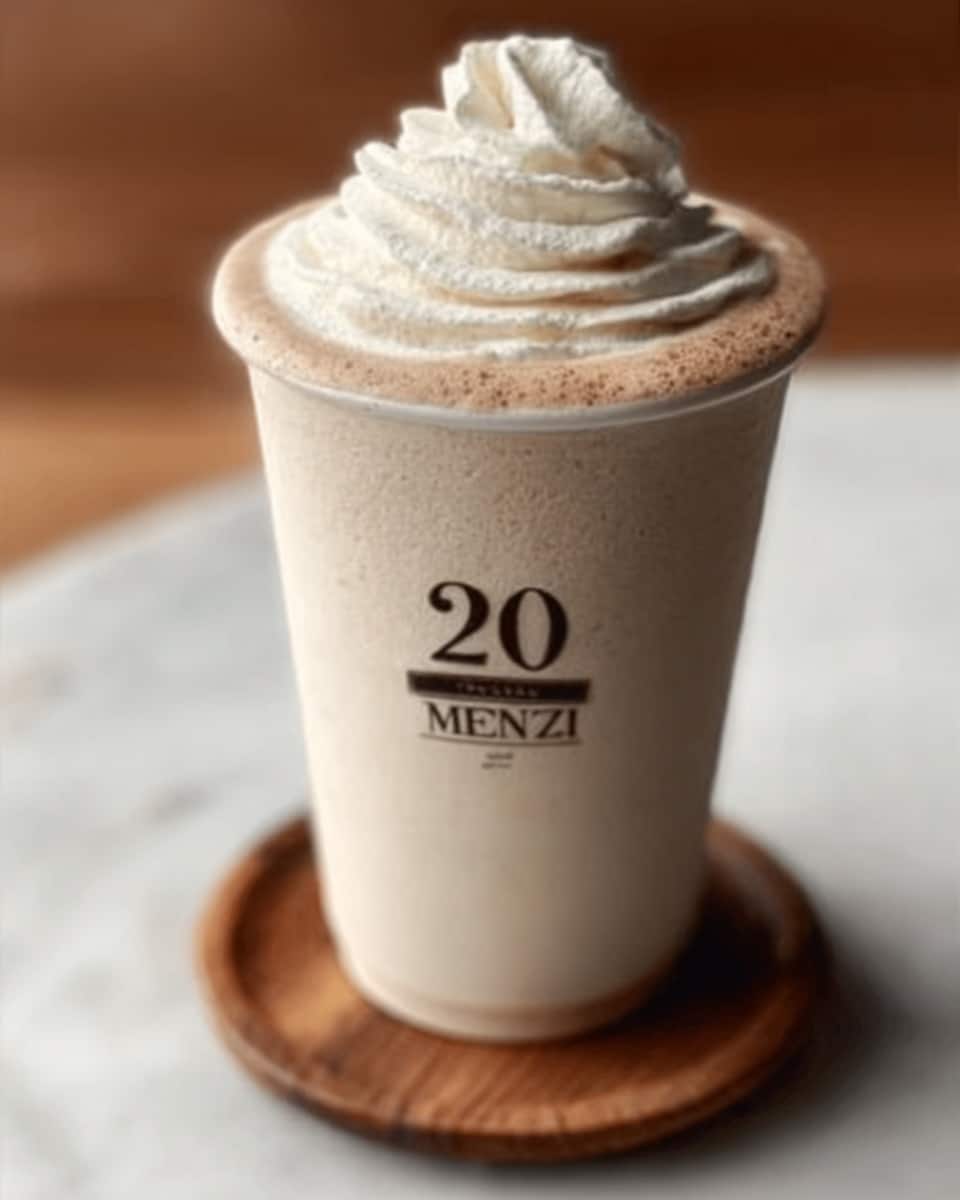 A tall white cup filled with a light brown creamy drink topped with a swirl of white whipped cream with a soft and smooth texture. The cup has simple black text that says