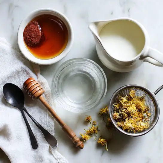 A flat lay image shows a clear glass cup in the center filled with water, surrounded by a white ceramic pitcher filled with milk on the upper right and a white small bowl containing dark amber honey on the left. Next to the honey, a wooden honey dipper lies horizontally, paired with a black spoon holding a reddish brown powder placed vertical along the left edge with a white cloth underneath. At the lower right, a metal tea strainer contains yellow dried flowers, with a few loose dried flowers scattered on the white marbled surface. photo taken with an iphone --ar 4:5 --v 7
