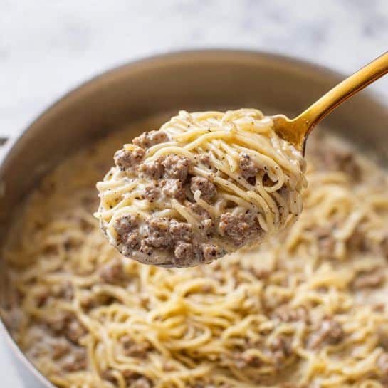 The image shows a white marbled surface with a clear glass bowl in the center filled with raw ground meat, dark red and pink in color. Below the bowl, there is a small stack of orange pasta. To the right of the meat bowl, there is a white bowl filled with sliced white and light brown mushrooms. Above the mushroom bowl, whole mushrooms are scattered on the marbled surface. To the top right, a small bunch of green parsley leaves is placed. Next to the parsley, there is an orange onion. Above the onion, a small white bowl with a yellow liquid, likely oil, is visible. To the left of the oil bowl, a small white bowl with a creamy white substance is present. Next to it, another small white bowl contains a pale yellow liquid, possibly broth or vinegar. To the far left at the top, there is a small white cup filled with white liquid, perhaps milk or cream, and beside it, a glass container with brown liquid, possibly broth. The photo taken with an iphone --ar 4:5 --v 7