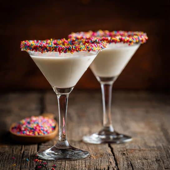 The image shows two clear martini glasses filled with a smooth white drink. The rims of the glasses are coated with a colorful mix of small candy sprinkles that add a bright touch. The glasses are standing on a rough wooden surface that contrasts with the clean look of the drinks. The texture of the drink looks creamy and thick, filling the glasses almost to the top. The background is softly blurred with warm dark brown tones, making the glasses stand out clearly. photo taken with an iphone --ar 4:5 --v 7