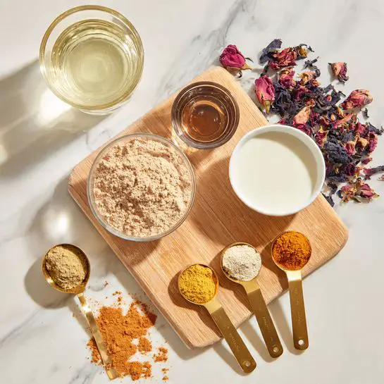 The image shows a light wooden cutting board on a white marbled surface with several ingredients arranged on and around it. On the board’s top left, there is a small clear glass bowl filled with a light brown powder. Next to it, to the right, is a white bowl filled with white liquid. Below the powder bowl is a small glass container with a dark brown liquid inside. On the right edge of the board, three gold measuring spoons are lined up, each holding a different powdered spice: an orange-yellow powder, a light beige powder, and a brown powder. A gold spoon with similar light brown powder sits on the marbled surface to the left of the board. Near the top right corner of the board, scattered dried dark purple and pink flower petals add a touch of color. A clear empty glass is positioned at the top left corner of the image. photo taken with an iphone --ar 4:5 --v 7