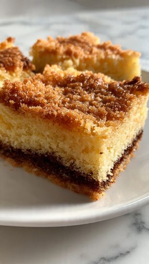 A single slice of layered cake sits on a white plate, showing three layers of light brown cake separated by two layers of creamy frosting around the same color. The top layer is thick with pale yellow frosting and sprinkled with small brown crunchy bits. The cake looks moist and soft, and the plate is placed on a white marbled surface. In the background, the rest of the cake on a white stand is slightly out of focus. A woman's hand is holding the cake slice from the side. Photo taken with an iphone --ar 4:5 --v 7