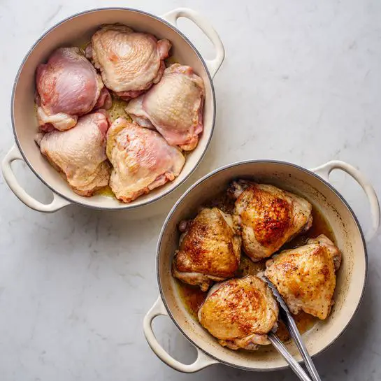 The image shows two white pots on a white marbled surface. The top pot has five raw chicken thighs, pink with white fat parts and some seasoning, sitting in light oil. The chicken pieces are arranged close but not overlapping, showing a mix of smooth and slightly bumpy textures. The bottom pot contains four cooked chicken thighs with a crispy golden-brown skin that looks crunchy, sitting in browned oil with some cooking marks visible on the pot's sides. A pair of silver tongs is holding one of the cooked pieces. Both pots have large handles and a light cream interior. Photo taken with an iphone --ar 4:5 --v 7