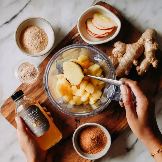 A woman's hand is holding a spoon over a clear blender container filled with orange chunks and a large slice of yellow ginger. The blender container is on a wooden surface. Below the container, another woman's hand holds a large bottle of light amber liquid labeled as apple cider vinegar. Surrounding the blender on the wooden surface are a piece of ginger root, a small white bowl with two slices of apple, a larger white bowl filled with light brown sugar, and a small white bowl with a reddish-brown powder. The background is a white marbled texture. photo taken with an iphone --ar 4:5 --v 7