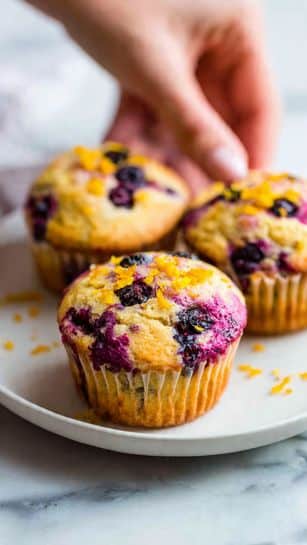 The image shows several muffins with crumbly, golden-yellow tops mixed with red spots of cranberry, scattered on a white marbled surface. The tops look soft with bits of crumbly texture, and the muffins themselves are light brown at the bottom with a baking paper liner seen on one of them. Around the muffins are bright red whole cranberries and a few small orange peel curls, adding contrast and color to the scene. There is a white bowl filled with shiny red cranberries placed among the muffins, creating a fresh and inviting look. Photo taken with an iphone --ar 4:5 --v 7