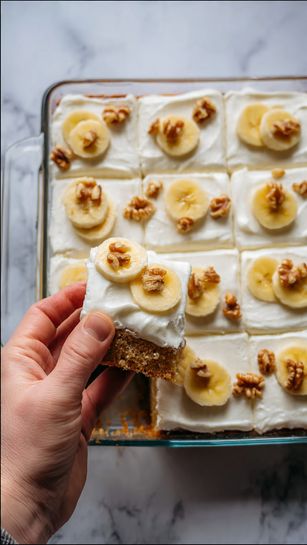 The image shows a rectangular glass baking dish filled with a cake cut into 16 square pieces. The top layer is thick white frosting, smooth and creamy, each square decorated with a slice of banana and small walnut pieces. The cake layer beneath is visible in the corner where a piece is removed, showing a light brown, moist texture. The dish sits on a white marbled surface, and a woman's hand is holding the removed piece. photo taken with an iphone --ar 4:5 --v 7