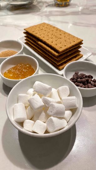 A white bowl full of small white marshmallows sits on a white marbled surface next to a white plate with stacked rectangular graham crackers. Nearby are three small white bowls containing brown sugar, orange honey, and dark brown chocolate chips. photo taken with an iphone --ar 4:5 --v 7