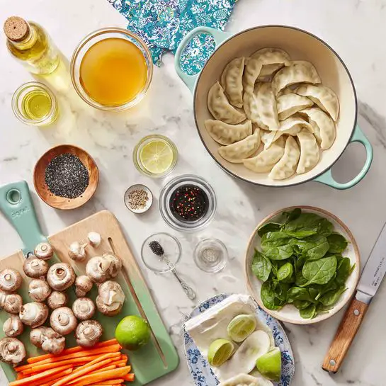 A white enameled pot with light green handles sits at the top right on a white marbled surface, surrounded by various ingredients for cooking. Below the pot, a beige plate holds semi-circular dumplings arranged in a circular pattern. To the right of the dumplings, a small metal bowl is filled with fresh spinach leaves. In the center is a glass measuring cup containing light orange broth. Above it is a small glass bowl with dark chili oil, next to a small wooden plate with black sesame seeds and a blue measuring spoon. A bottle of light yellow oil is located at the top left near a blue patterned cloth. To the left bottom, thin orange carrot strips are in a white bowl stacked on a larger white plate with blue rims. Below that, a green cutting board holds various sliced and whole brown mushrooms, two whole limes and one halved lime. Nearby, three garlic cloves and two green onions with white bulbs lie beside a knife with a wooden handle. Light green measuring cups are stacked near the cutting board. The whole scene is bright and neatly arranged. photo taken with an iphone --ar 4:5 --v 7