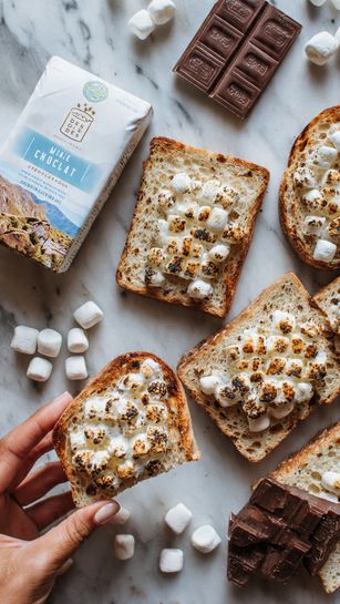 The image shows a close-up of several slices of toasted bread topped with small, white marshmallows, which are toasted to a light golden brown. The toast is placed on a white marbled surface alongside square pieces of dark chocolate and a white package labeled milk chocolate with blue and brown accents. The arrangement includes whole marshmallows scattered around, giving a cozy, sweet snack feel. A woman's hand holds one piece of the toast, highlighting the soft texture of the marshmallows and the crunchy bread beneath. Photo taken with an iphone --ar 4:5 --v 7
