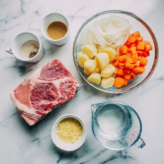 The image shows fresh ingredients arranged on a white marbled surface. There is a clear glass bowl with a thick piece of red meat with white fat marbled inside, placed on the right side. Above it, a large silver bowl holds chopped orange carrots, white onion slices, and yellow small potato pieces all layered side by side. To the left of the meat, there are two small white ramekins, one filled with a brown sauce and the other with a dry seasoning mix in light beige color. Below these, a small white dish contains a yellow minced substance. In the bottom right corner, there is a clear glass measuring cup filled with water. Everything is neatly placed and well-lit, giving a fresh and clean look. photo taken with an iphone --ar 4:5 --v 7