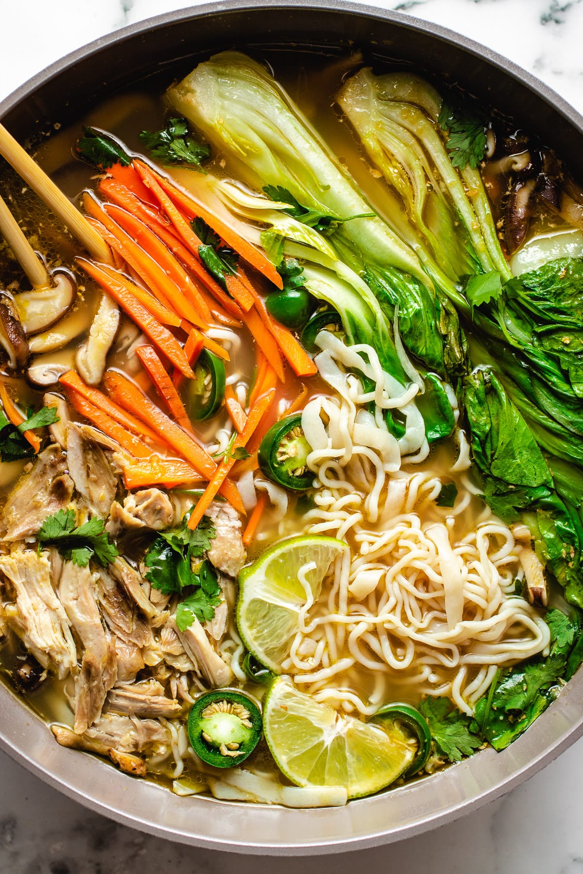 The image shows a bowl of soup with five visible layers and colorful ingredients arranged neatly. At the bottom, there are light yellow noodles in a clear, golden broth. On the right side, there are green leafy bok choy with a smooth, shiny texture. Next to the bok choy are slices of bright green jalapeño peppers. On the left side, thin orange carrot sticks are placed vertically, with some fresh green herbs sprinkled on top. Below the carrot sticks, shredded cooked chicken pieces sit lightly scattered. Two slices of lime rest on the noodles near the jalapeños, adding a fresh touch. The bowl is white and placed on a white marbled surface. A pair of wooden chopsticks rests on the left side of the bowl. Photo taken with an iphone --ar 4:5 --v 7
