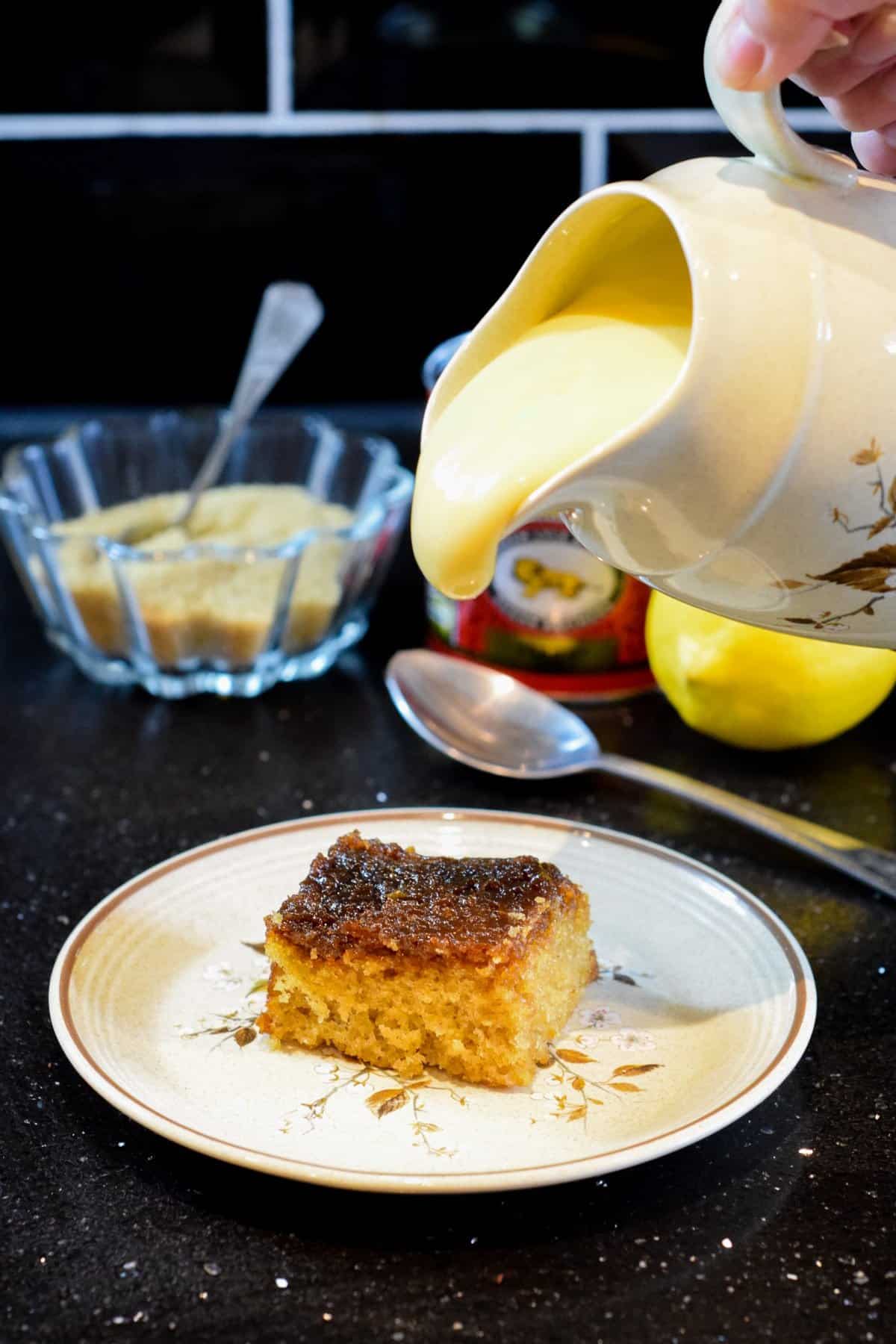 A small round cake with two visible layers sits on a white plate with a floral design. The top layer is a darker golden brown with a slightly shiny and crumbly texture, while the bottom layer is lighter brown and soft with a crumbly interior. A wedge has been removed, showing the moist and grainy inside. There are a few crumbs scattered on the plate around the cake. The background features green leaves and a white marbled surface. Photo taken with an iphone --ar 4:5 --v 7