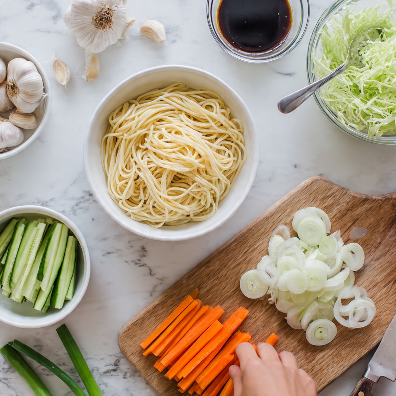 The image shows a top view of a white bowl filled with cooked noodles on the left side, with pale yellow and smooth texture. Below it, there is a white bowl with sliced cucumber sticks, long and green, visible on the lower left corner. In the center, a wooden board holds orange carrot sticks, sliced thick and arranged neatly. Next to the carrots, there are sliced white onions with a slightly translucent texture, loosely placed. Above the onions is a clear glass bowl with light green shredded cabbage. On the upper right is a white bowl with a dark soy sauce and a silver spoon resting inside. Scattered around are whole garlic cloves, a rusty cleaver knife with a wooden handle, and green scallions on a white marbled surface. A woman's hand is reaching towards the bowls. Photo taken with an iphone --ar 4:5 --v 7