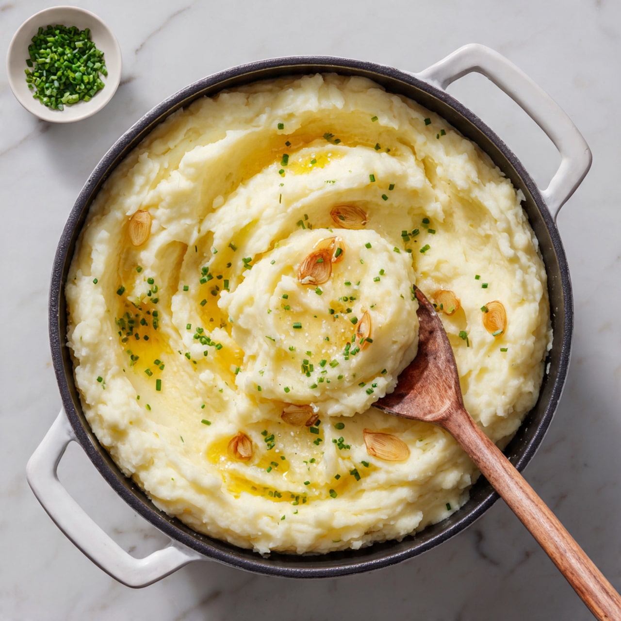 The image shows a pot filled with creamy mashed potatoes that have a soft and smooth texture with some small lumps. The mashed potatoes are light yellow with a swirl of melted butter on top, creating a shiny golden layer. Roasted garlic cloves with a light brown color are scattered on the surface, along with small green chive pieces sprinkled evenly. A wooden spoon is scooping some mashed potatoes from the center, showing a dense and creamy inside. The pot is black with white handles, and it rests on a white marbled surface. In the background, there is a small white bowl with chopped chives. Photo taken with an iphone --ar 4:5 --v 7