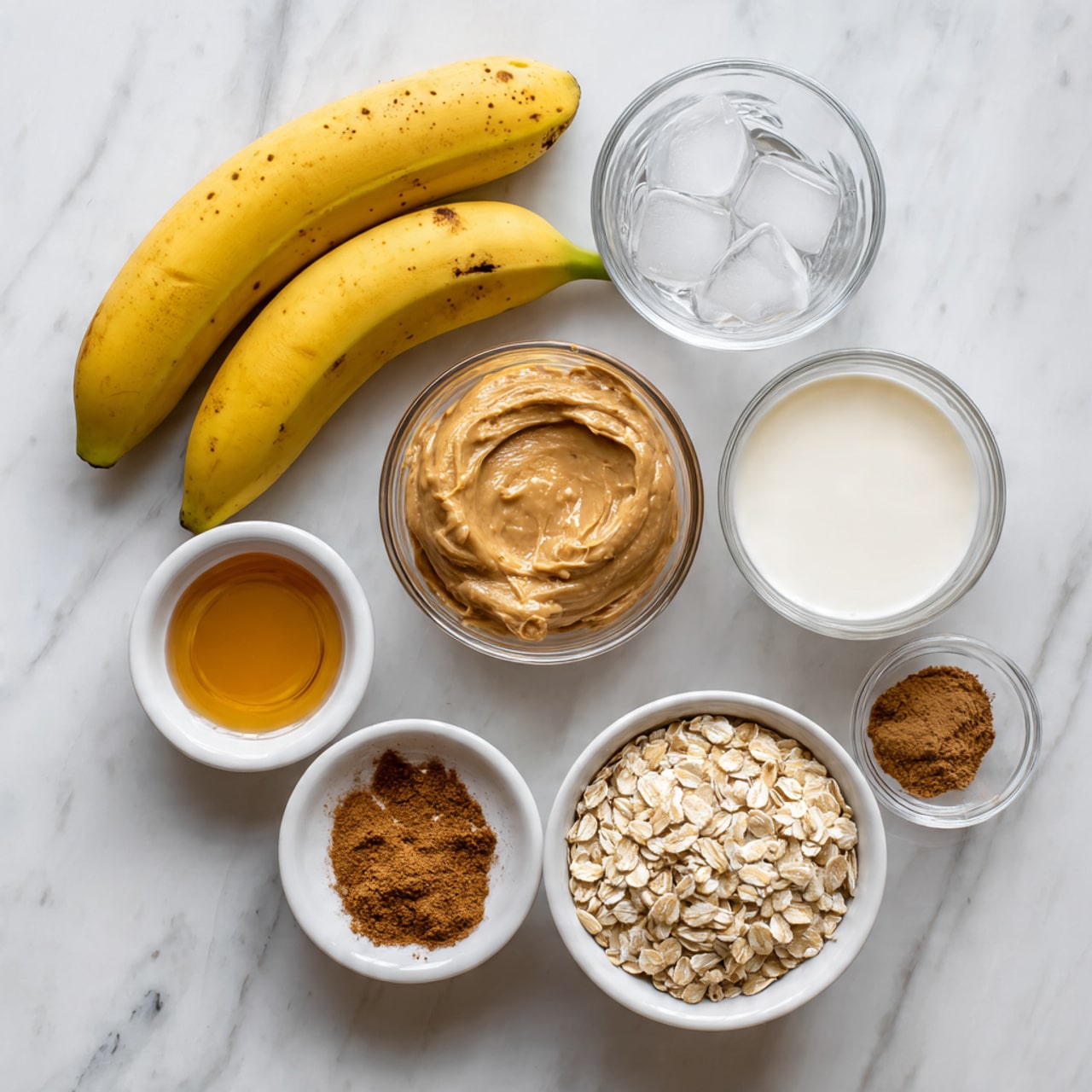 The image shows ingredients neatly placed on a white marbled surface. At the top left, there are two whole yellow bananas with slight brown spots. On the right side, there is a small clear glass of water with four ice cubes and below it a small white bowl filled with a creamy white substance. In the center, a clear glass bowl holds a smooth light brown peanut butter spread. Below that, a white bowl is full of rolled oats. To the bottom left, there is a small white bowl with a golden brown liquid and next to it another small white bowl containing ground brown cinnamon. Above this is a clear glass bowl with a white liquid, likely milk, positioned slightly above and to the right of the peanut butter bowl. photo taken with an iphone --ar 4:5 --v 7