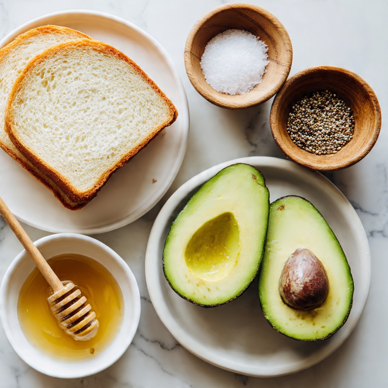 The image shows a simple arrangement of food items on a white marbled surface. There is a slice of white bread on a white plate with a light golden crust and soft texture. Next to the bread are two halves of a fresh avocado with bright green flesh and a brown seed in one half. Below the avocado, there is a white bowl filled with golden honey, with a wooden honey dipper resting inside. Above the bread and avocado, there are two small wooden bowls, one filled with coarse salt and the other with black peppercorns. The lighting is soft and natural, highlighting the colors and textures of each item. Photo taken with an iphone --ar 4:5 --v 7