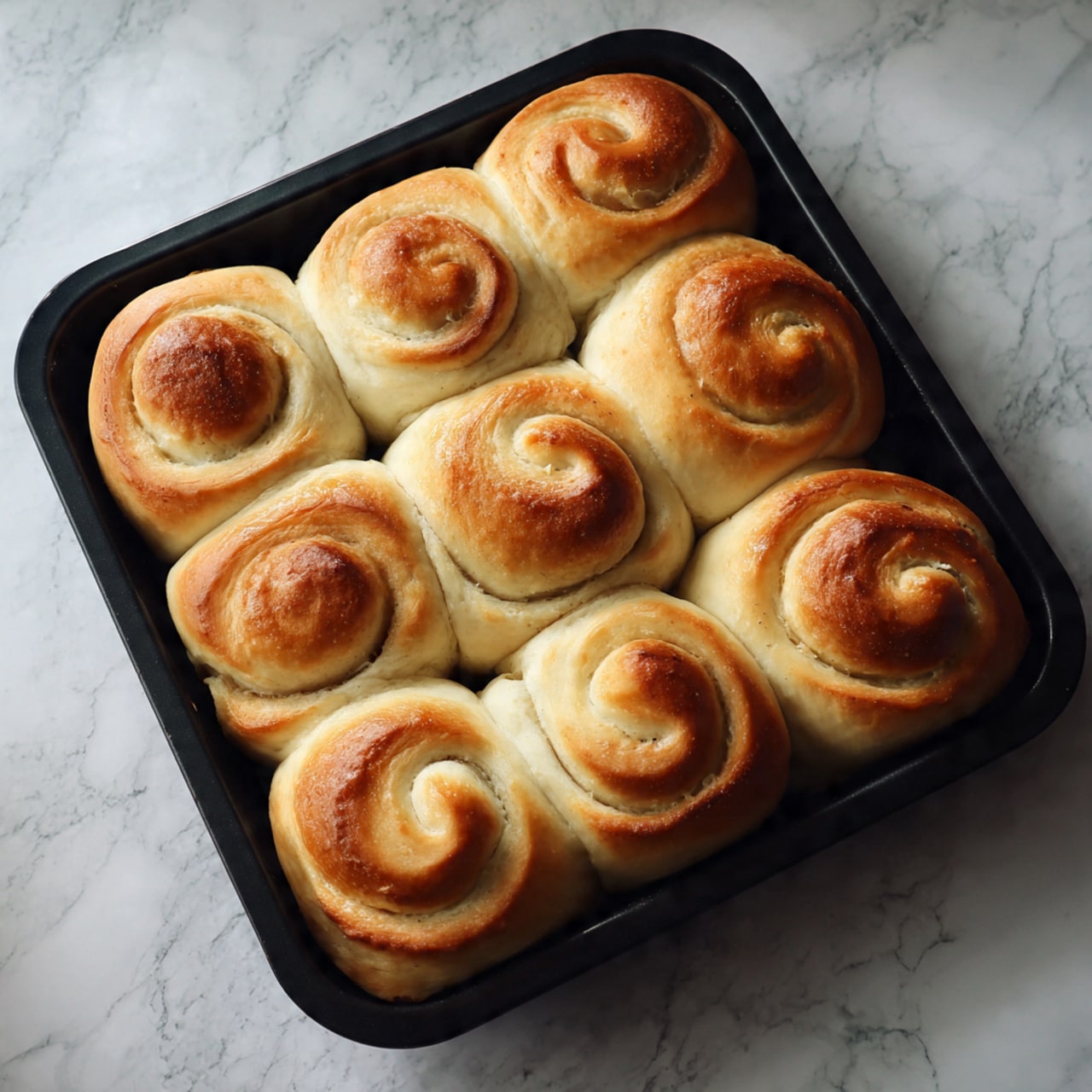The image shows a black baking tray filled with about twelve spiral-shaped rolls of dough, each with a golden-brown top and lighter sides. The rolls are arranged close to each other in rows, with a soft and slightly puffy texture visible on the dough. The background surface is a white marbled texture. Photo taken with an iphone --ar 4:5 --v 7