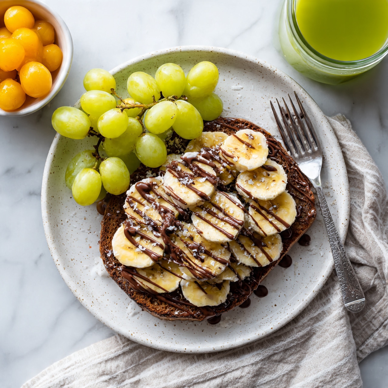 A white speckled plate holds one large round dark brown toast, topped with many slices of pale yellow banana covering the entire toast in one layer. Over the bananas, thin stripes of dark chocolate spread are drizzled in a zigzag pattern. To the upper left of the plate, there is a small white bowl filled with bright orange and pale green grapes. To the right behind the plate, a transparent glass is filled with a light greenish drink. A woman's hand with a fork is near the plate, resting on a folded, light beige and white striped napkin on a white marbled surface photo taken with an iphone --ar 4:5 --v 7