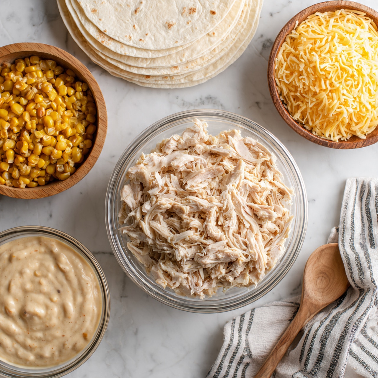 The image shows a white marbled surface with several clear glass bowls arranged on it. In the middle, there is a bowl filled with shredded cooked chicken, light beige in color with soft texture. To the top left, a stack of white tortillas sits neatly. On the right side, a bowl is filled with shredded yellow cheese, finely grated and fluffy. Above the chicken bowl, there is a small bowl holding cooked corn kernels, yellow with some brown spots. At the bottom left, a bowl contains a thick creamy sauce with a smooth texture in a light beige color. On the right side of the image, a striped cloth napkin is partially visible with a wooden spoon resting on it. Photo taken with an iphone --ar 4:5 --v 7