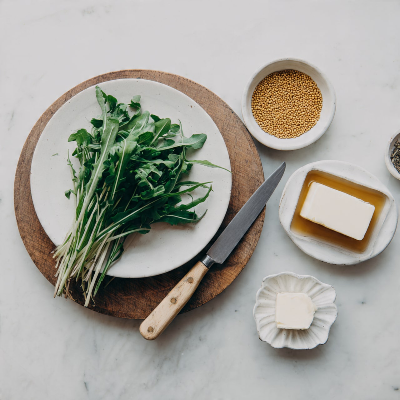 A white plate holds a round wooden board topped with a small pile of fresh green herbs with thin stems and leaves, placed near the left side. To the right of the plate is a medium-sized knife with a light wooden handle and a silver blade resting on a white marbled surface. Above, there are three small dishes: a round white bowl filled with coarse yellow mustard seeds placed near the top center, a square white dish with a golden-brown sauce beside it to the right, and a small white flower-shaped bowl containing a pale square slab of butter below the square dish. The overall setting is on a white marbled surface, giving a clean and simple background. Photo taken with an iphone --ar 4:5 --v 7