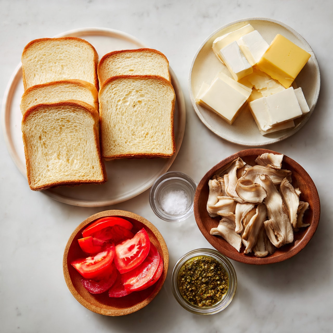The image shows sandwich ingredients arranged on a white marbled surface. On the left, there is a white plate with five slices of golden brown bread, showing a soft inside texture. To the right, a wooden bowl holds several thin slices of light beige cooked meat. Above the meat, a clear plate contains square slices of pale yellow cheese. Next to the cheese, a small clear bowl holds a few white butter cubes. Below the bread, a wooden bowl has bright red, thin tomato slices. At the bottom center, a small clear bowl contains a green, coarse sauce, likely pesto. photo taken with an iphone --ar 4:5 --v 7