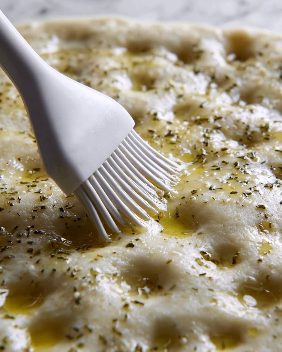 A close-up image of dough covered with small holes and shiny pools of olive oil sitting in the dimples. The dough is light beige with a soft and fluffy texture, speckled with small dried herbs. A white silicone brush with soft bristles is gently spreading the oil and herbs mixture on the dough, creating a glossy surface. The background is a white marbled texture. Photo taken with an iphone --ar 4:5 --v 7