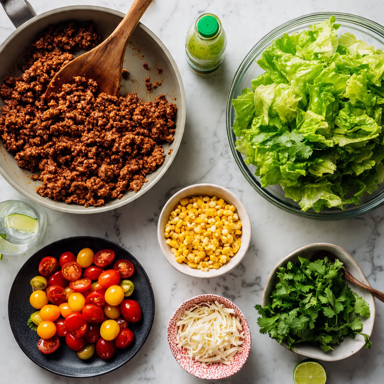 Taco Salad Jars Recipe 4 The image shows a top-down view of ingredients laid out on a white marbled surface. There is a large grey pan at the top left filled with cooked ground meat, and a wooden spoon resting inside it. Next to the pan on the right is a clear glass bowl filled with fresh green lettuce leaves. A bottle of green-capped dressing is placed above the bowl. Below the pan, there is a small black plate filled with halved cherry tomatoes in red and yellow colors. To the right of the tomatoes, there is a small pink patterned bowl with yellow corn kernels, and next to that, a small white bowl filled with shredded white cheese. At the bottom, there is a small white bowl with fresh green cilantro leaves. There is also a clear glass with lime water positioned near the cilantro bowl. The photo taken with an iphone --ar 4:5 --v 7
