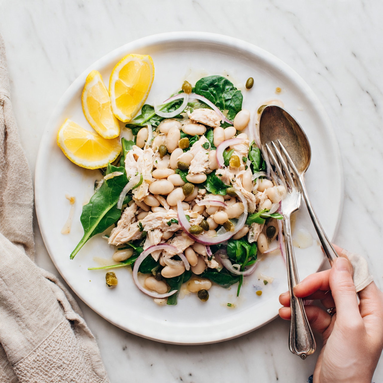 A white plate sits on a white marbled surface, filled with a fresh salad showing three main layers: the bottom layer of large, dark green spinach leaves, the middle layer of white beans and sliced green olives, and the top layer of cherry tomato halves, light pink tuna chunks, and thin red onion slices mixed with fresh chopped green herbs and parsley leaves scattered around. A vintage silver fork rests on the right side of the plate, slightly inside the salad. In the background, there is a blurred amber glass with a textured pattern and some fresh green herbs at the edges of the frame. photo taken with an iphone --ar 4:5 --v 7