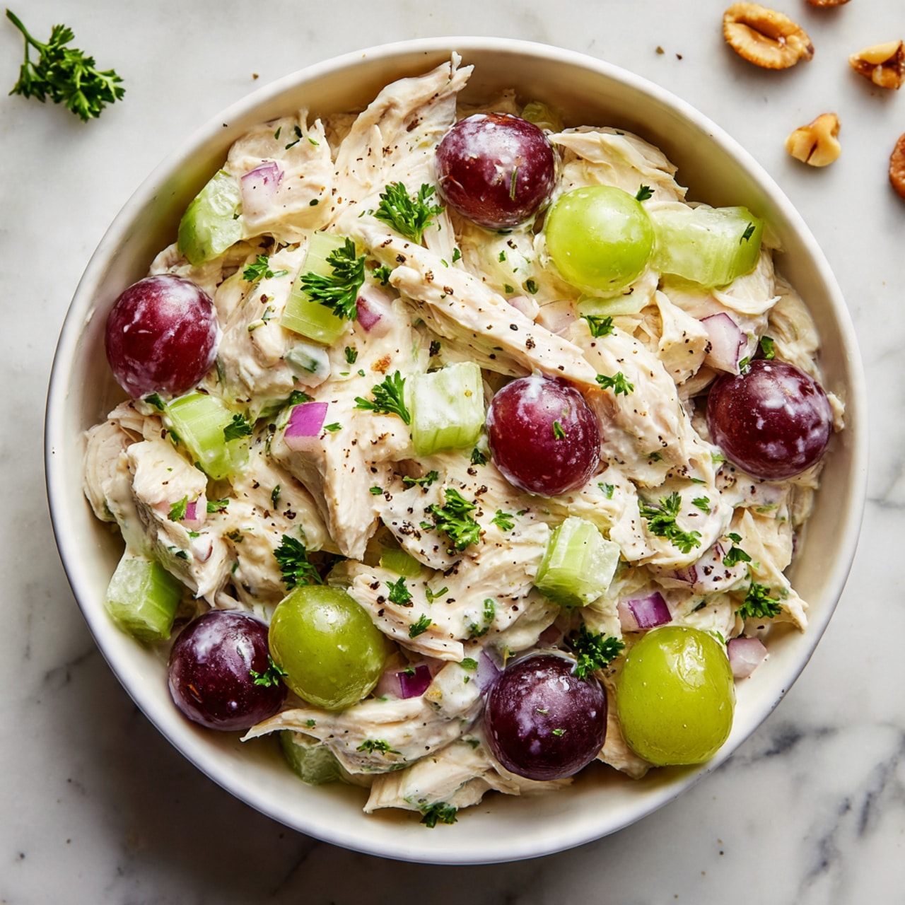 A white bowl filled with a creamy chicken salad that has three main layers: shredded white chicken mixed with a light creamy dressing forms the base layer, then there are sliced green celery pieces with a crisp texture mixed throughout, and halved deep purple-red grapes scattered evenly on top. Small chunks of finely chopped purple onion are mixed throughout, adding pops of color, while fresh green parsley leaves are sprinkled on top for garnish. A light dusting of black pepper is visible on the creamy mix. The bowl sits on a white marbled surface with some small scattered pieces of nuts in the background. Photo taken with an iphone --ar 4:5 --v 7