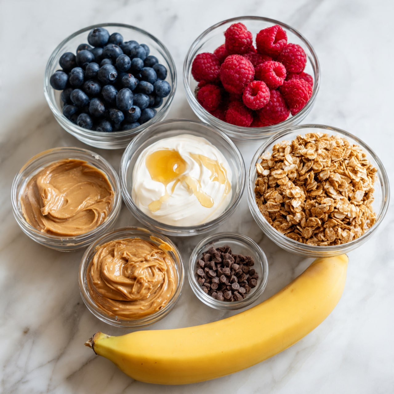 The image shows seven clear glass bowls of different sizes and a ripe yellow banana arranged on a white marbled surface. The largest bowls hold fresh blueberries and bright red raspberries, placed side by side near the top. Next to them is a bowl containing a creamy white substance with a drizzle of honey. Below, there are smaller bowls with light brown peanut butter, golden brown granola, and dark brown cacao nibs. The yellow banana lies to the right below the bowls, with smooth, bright skin and a light green stem. The overall color palette is fresh and natural with crisp details. Photo taken with an iphone --ar 4:5 --v 7