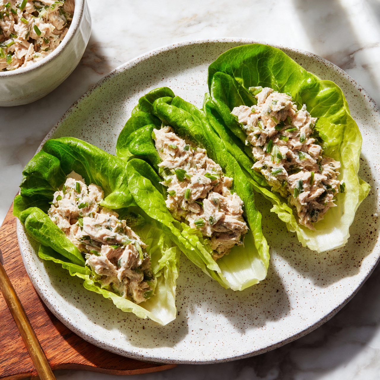 The image shows three bright green lettuce leaves placed in a row on a white speckled plate, each leaf filled with a creamy, light beige tuna salad with small dark green herbs mixed in. The lettuce leaves are smooth and fresh, and the tuna salad has a slightly chunky texture that fills the middle of each leaf. The plate sits on a white marbled surface with a wooden edge visible on one side, and a white bowl filled with more tuna salad is partly visible in the background. The scene is lit with soft natural light, highlighting the colors and textures clearly. photo taken with an iphone --ar 4:5 --v 7