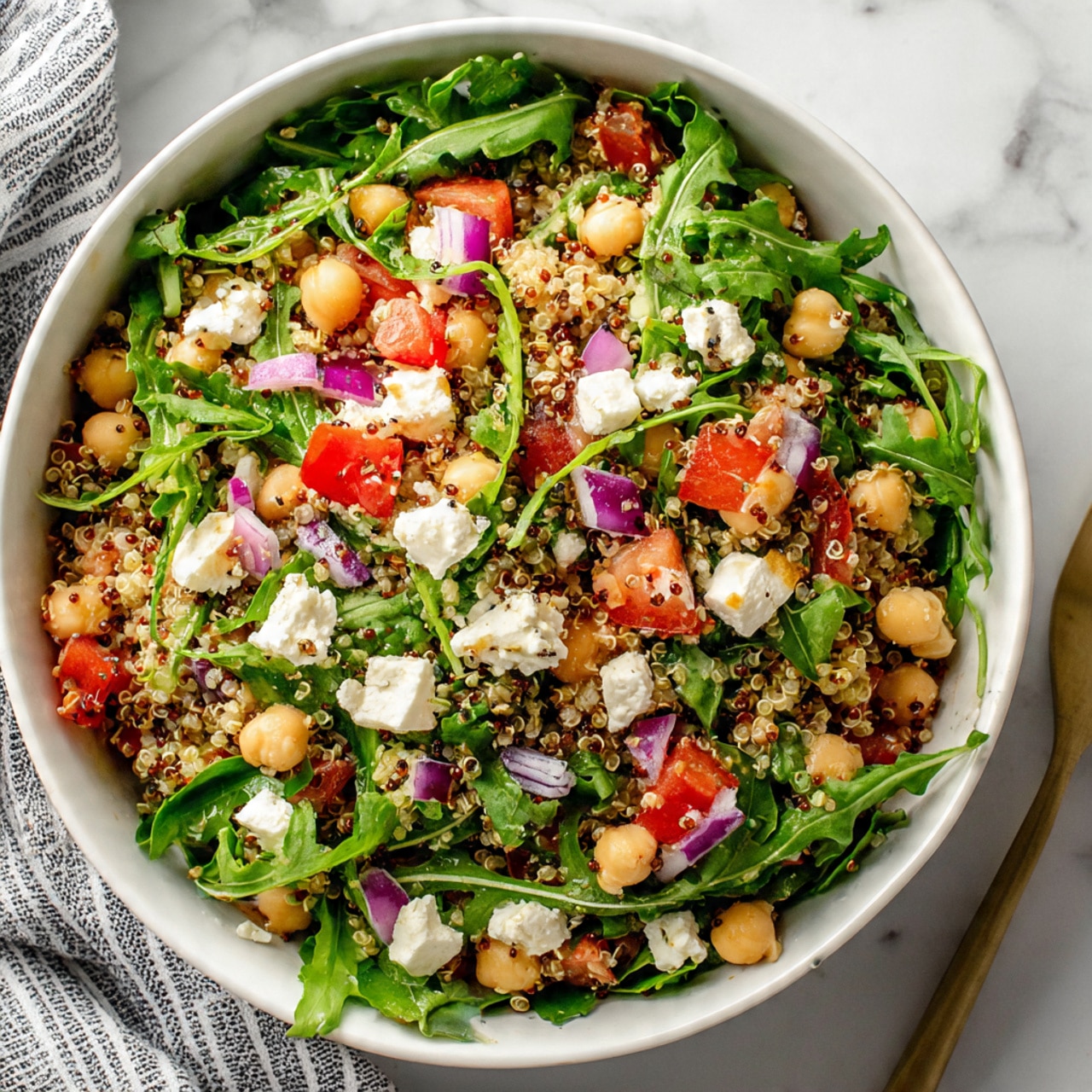 A white bowl filled with a fresh salad showing three main layers: the bottom layer is bright green leafy vegetables mostly arugula, the middle layer is a mix of small white chickpeas and light tan cooked quinoa grains, and the top layer has chopped red tomatoes and small bits of purple onion scattered around, adding spots of red and purple color. Small white cubes of cheese are mixed in evenly with the other ingredients, and the salad looks lightly seasoned with dark specks of pepper over it. The bowl is placed on a white marbled surface with a striped cloth nearby. Photo taken with an iphone --ar 4:5 --v 7