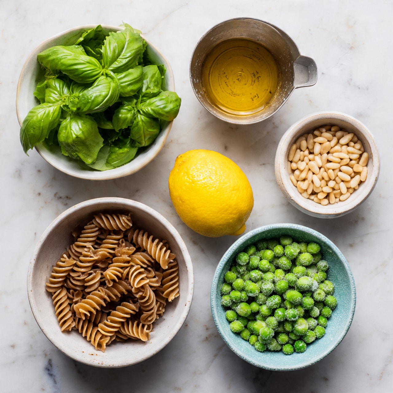 The image shows six small containers arranged on a white marbled surface with fresh ingredients for cooking. Starting from the top left, there is a white bowl filled with bright green fresh basil leaves with a smooth texture. On the top right, a metal measuring cup holds a small amount of golden brown liquid, likely oil. To the right middle, a small white bowl contains pale beige pine nuts, round and smooth. Below that, a halved bright yellow lemon sits with a visible juicy interior. On the left bottom, a white bowl holds dry brown spiral pasta with ridged texture. Lastly, at the bottom right, a white bowl with a slight blue tint around the rim is filled with frozen green peas, showing a frosty surface. Photo taken with an iphone --ar 4:5 --v 7
