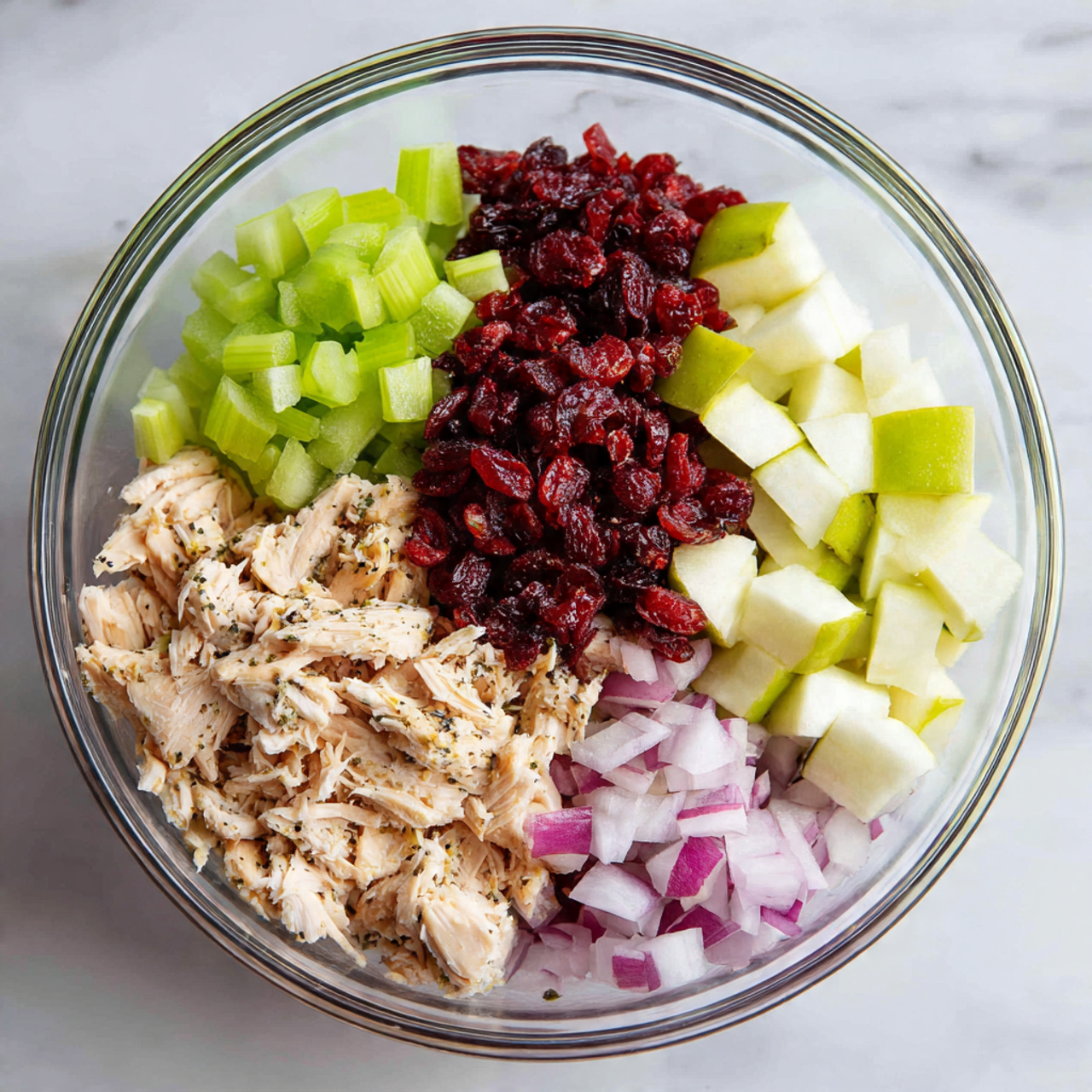A clear glass bowl sits on a white marbled surface, filled with five distinct layers. At the bottom left, there is a pile of shredded pale beige chicken with black specks of seasoning. Above it, to the left, are small cubes of bright green celery. In the center top, there is a heap of dark red dried cranberries with a slightly wrinkled texture. To the right of the cranberries, small white and light green apple cubes are stacked. Below the apple cubes and to the right of the chicken are small diced pieces of purple-red onion with white veins. The ingredients are neatly arranged without mixing, showcasing a variety of colors and textures. Photo taken with an iphone --ar 4:5 --v 7