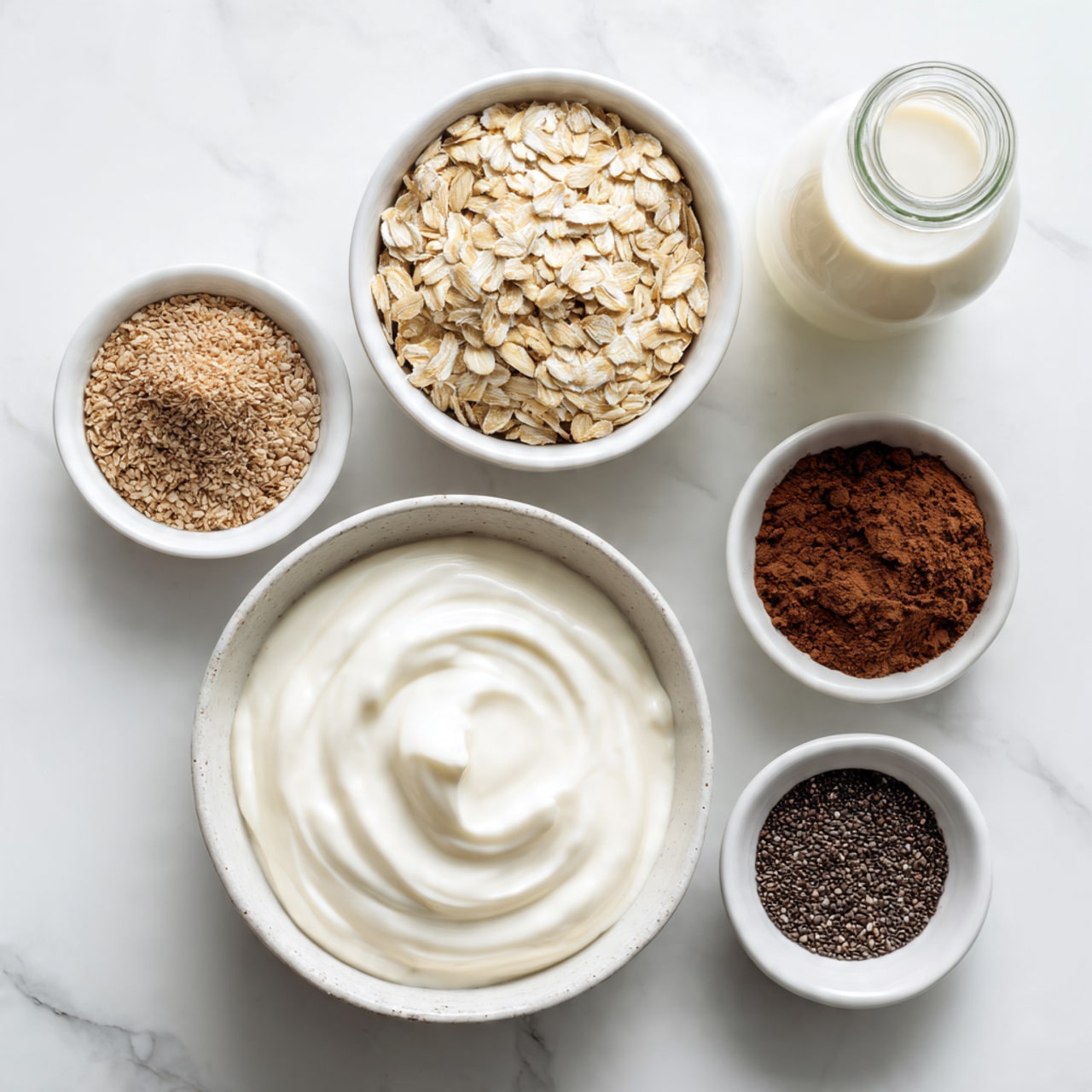 The image shows six white bowls and a glass bottle arranged on a white marbled surface. The largest bowl at the bottom center is filled with smooth, creamy white yogurt with a gentle swirl on top. Above it, a medium white bowl holds light brown rolled oats with a rough texture. To the left, there are two smaller white bowls, the top one containing light brown powder and the bottom one filled with tiny black chia seeds. On the right side, two more small white bowls hold darker brown cocoa powder and a small amount of dark liquid, likely vanilla or syrup. At the top right corner, a clear glass bottle contains white milk. photo taken with an iphone --ar 4:5 --v 7