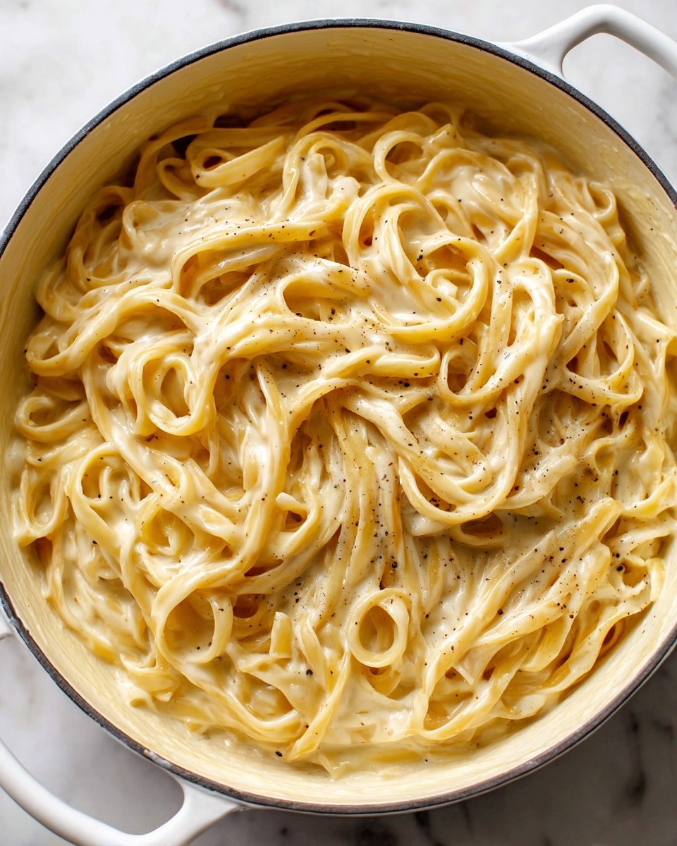 A close-up top view of a white pot filled with creamy fettuccine pasta. The pasta noodles are long, flat, and twisted together in thick clusters, coated evenly in a rich, smooth, light beige sauce with tiny black pepper specks spread throughout. The creamy sauce has a glossy texture, pooling slightly at the bottom edges of the pot. The pot rests on a white marbled surface, giving a clean and bright background. photo taken with an iphone --ar 4:5 --v 7