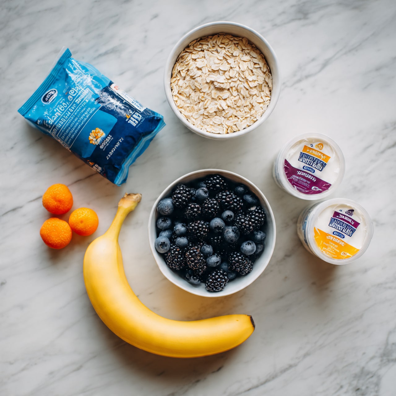 The image shows ingredients arranged on a white marbled surface, including a blue and white bag of oats positioned in the top left corner, a white cup filled with oats placed to the right of the bag, a white bowl full of blackberries and blueberries near the center, two small bright orange berries to the left of the white bowl, a yellow ripe banana curved along the bottom edge, and two white containers of yogurt with colorful labels resting between the bag of oats and the fruit. photo taken with an iphone --ar 4:5 --v 7