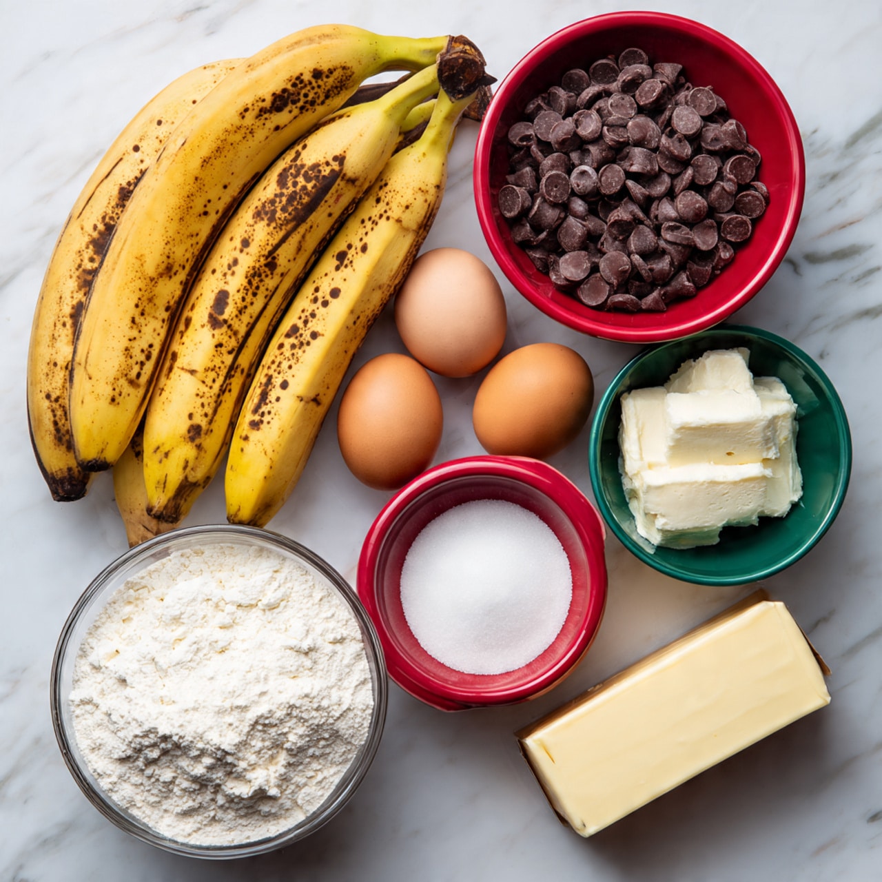 The image shows ripe bananas with brown spots positioned diagonally on the top left, two brown eggs next to them, and a white bowl filled with dark chocolate chips placed in the top right. Below the eggs, there is a red bowl filled with white granulated sugar and a small red bowl holding a white powdery substance next to it. To the right of the sugar bowl is a small green bowl with another white powder, likely baking soda or salt. In the bottom left corner, a clear glass bowl is filled with white flour, and at the bottom right corner, there is a stick of butter with yellow packaging. A small brown bottle of vanilla extract is near the sugar bowl, all placed on a white marbled surface photo taken with an iphone --ar 4:5 --v 7
