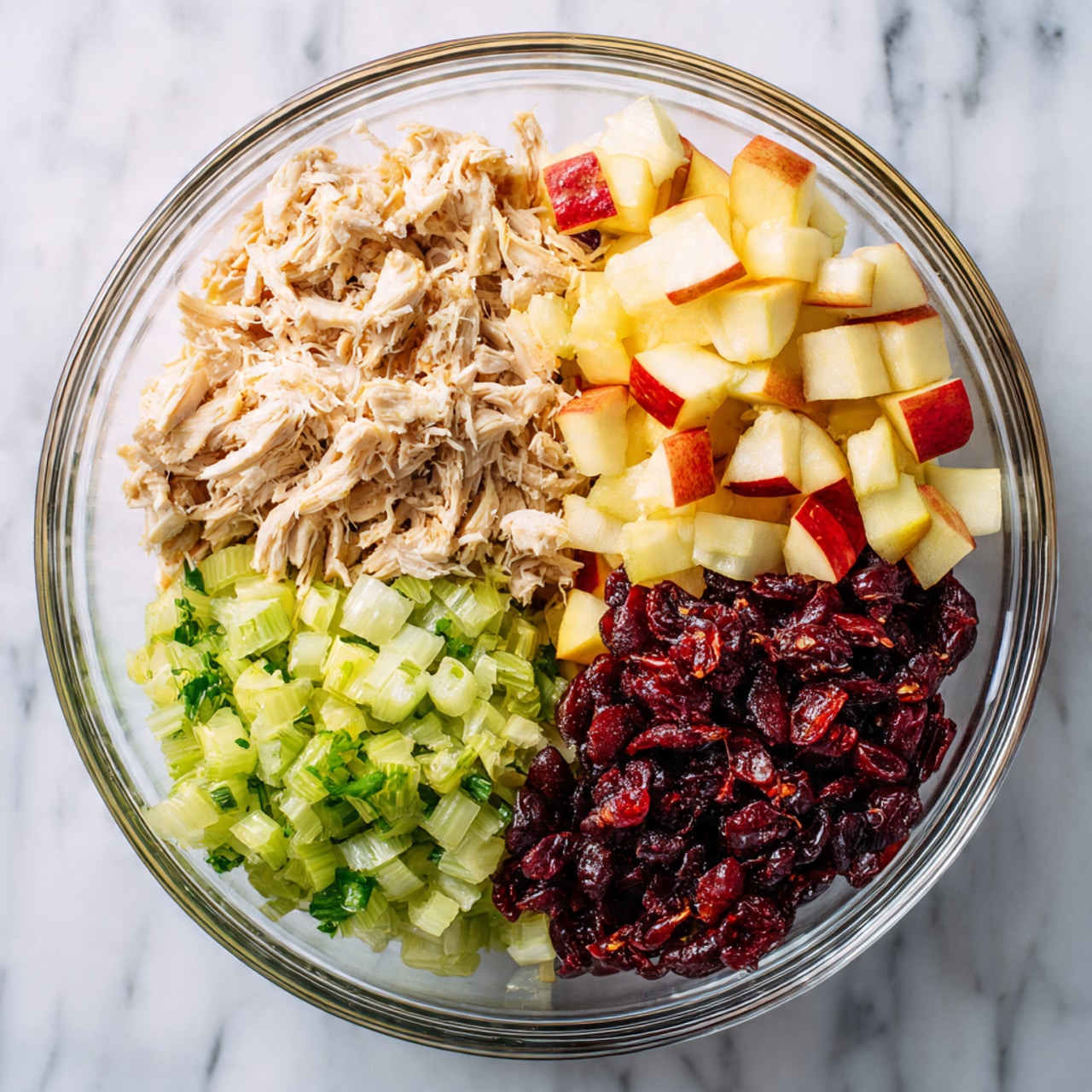 A clear glass bowl shows four separate layers of chopped ingredients. On the top left, there is light beige shredded chicken with a soft texture. On the top right, small diced pieces of red and yellow apple add a bright color. On the bottom left, finely chopped green celery pieces bring a fresh look. On the bottom right, a pile of dark red dried cranberries creates a rich color contrast. The bowl sits on a white marbled surface. Photo taken with an iphone --ar 4:5 --v 7