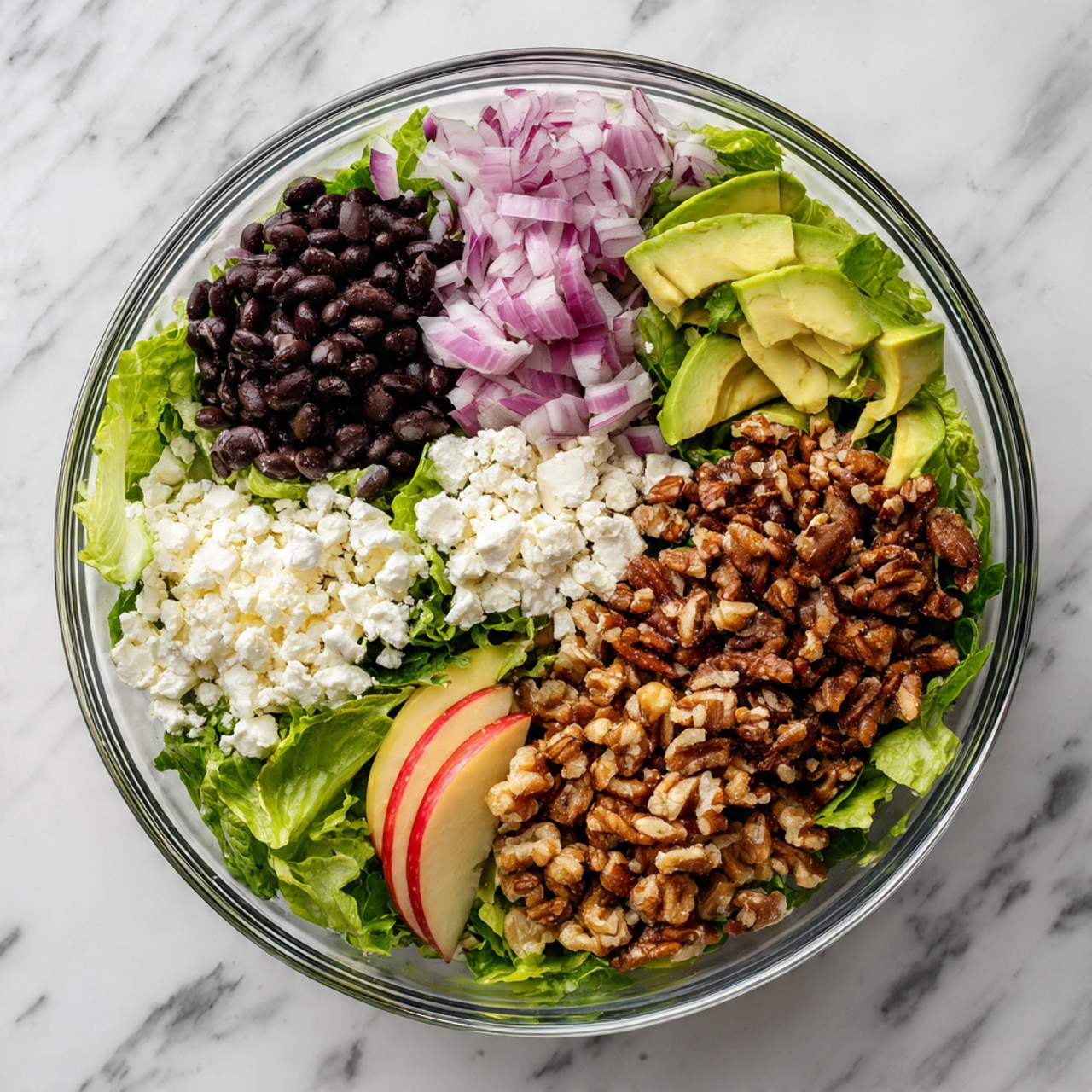 A clear glass bowl filled with a salad arranged in separate sections. At the bottom is a bed of green leafy lettuce. On the left side, there are dark black beans, next to white crumbled cheese. Above that, there are sliced purple onions, and next to the onions, light green avocado slices. On the right side, there is a large pile of brown chopped walnuts. At the bottom, there is a quarter of a red apple with its skin intact. The bowl is on a white marbled surface. Photo taken with an iphone --ar 4:5 --v 7